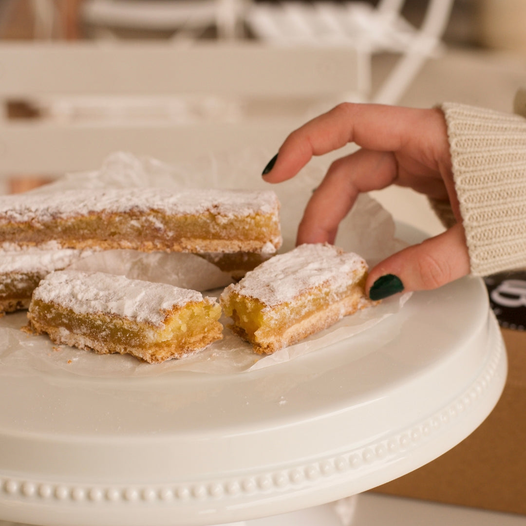 3 lemon bars sitting on a white cake plate. A woman's hand is picking up a piece that is cut in half.